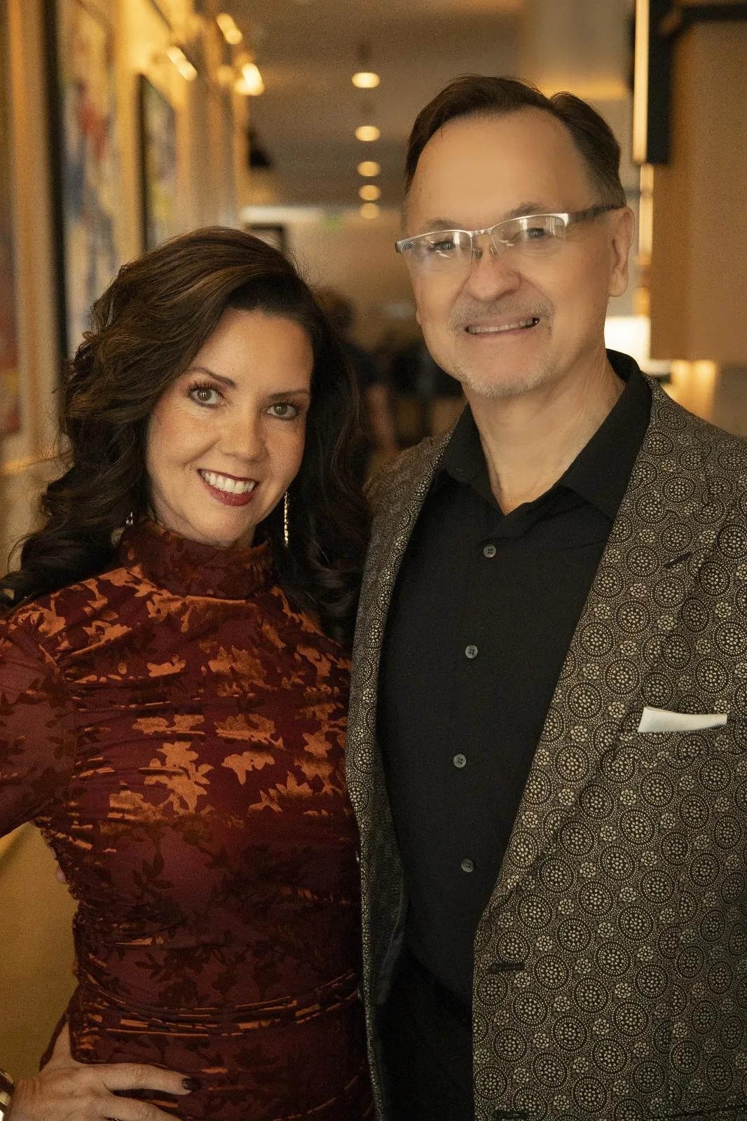 A woman in a patterned red dress and a man in a black shirt and patterned blazer stand side by side indoors, both smiling at the camera.
