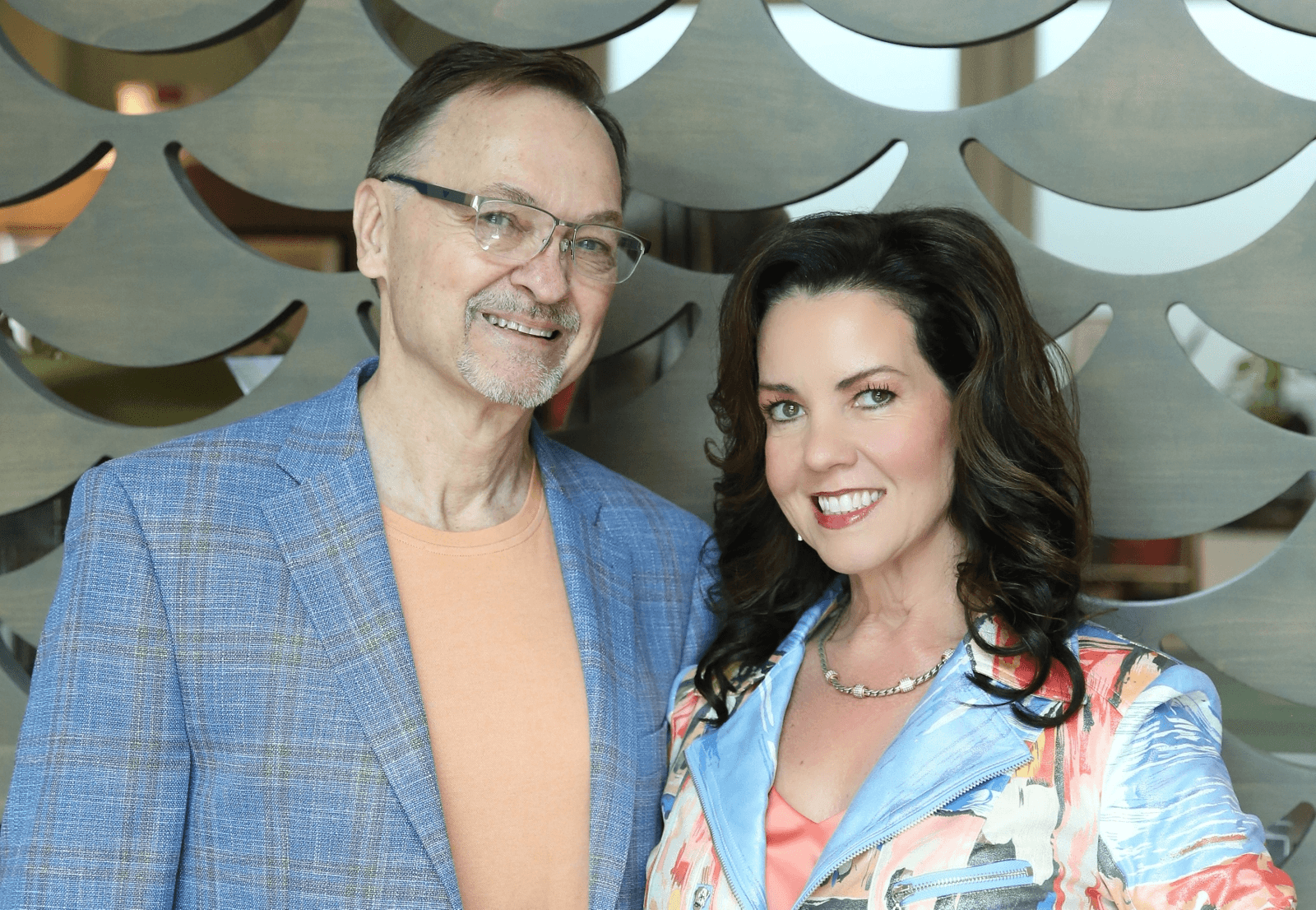 A man in a blue plaid blazer and an orange shirt stands next to a woman in a floral jacket, both smiling at the camera, with a decorative wooden backdrop behind them.