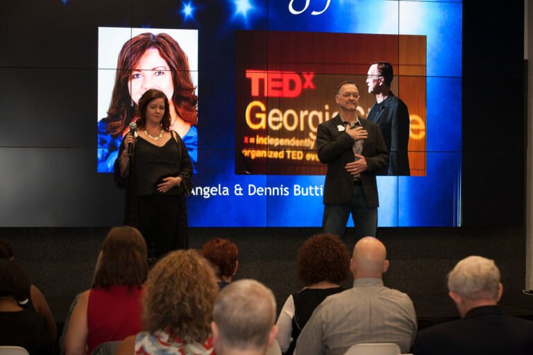 Angela and Dennis Buttimer stand on a tedx stage in front of an audience, with a presentation screen displaying their names in the background.