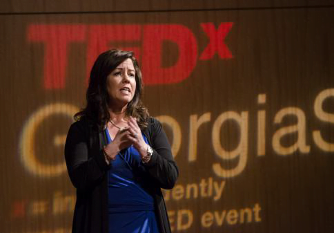 Speaker Angela Buttimer gesturing with her hands while presenting at a tedx event with the tedx logo and "georgiast" on the screen behind her.