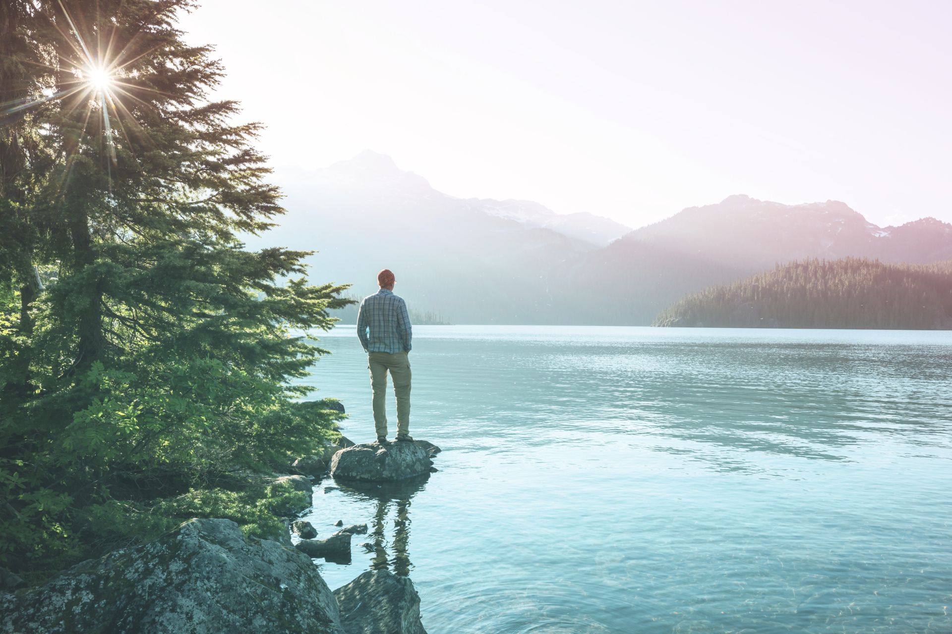 A monk stands on a rock at the edge of a serene lake surrounded by mountains and trees, with sunlight piercing through the sky.