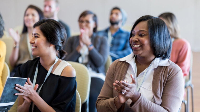 A group of people sitting in chairs, clapping, and smiling at an event exudes an air of refinement. In the foreground, two women are visible; one is holding a notebook.