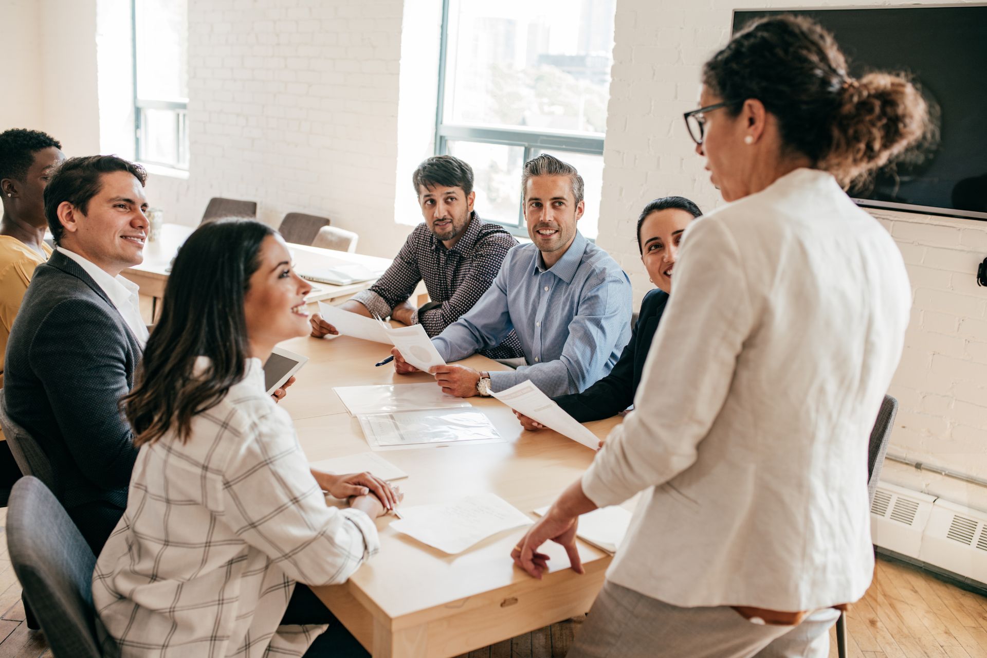 A group of professionals engaged in a discussion around a table while one person, demonstrating effective leadership, stands presenting a document.