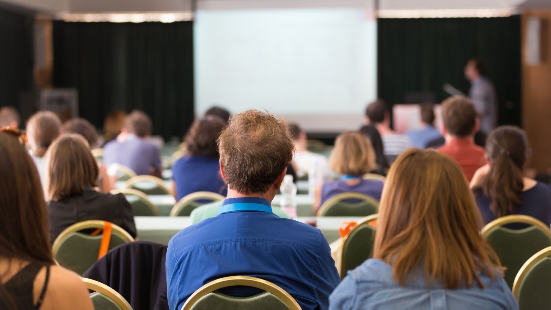 Audience in a conference room watches a presenter at the front near a projected screen during a training session.