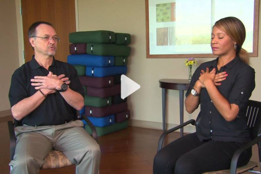 Two people practicing a breathing exercise, seated indoors with crossed arms on chests, a stack of yoga mats and a plant in the background, captured for media publication.