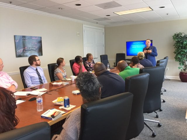 A group of professionals attentively listening to a presenter in a corporate meeting room with a digital slide visible in the background.