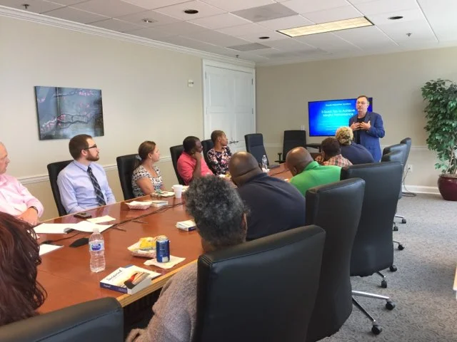 A group of professionals attentively listening to a presenter in a corporate meeting room with a digital slide visible in the background.