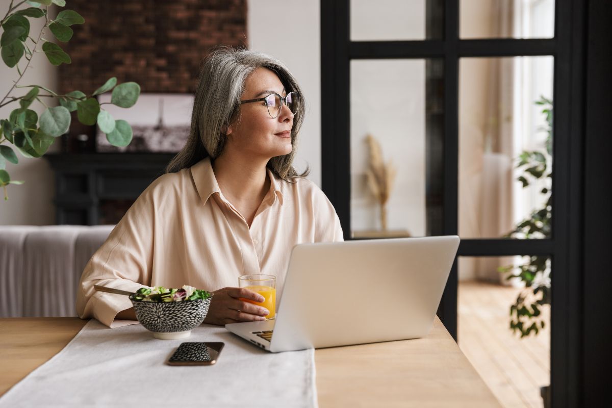 A woman in monk mode sits at a table with a laptop, holding a glass of juice, and looks out a window.