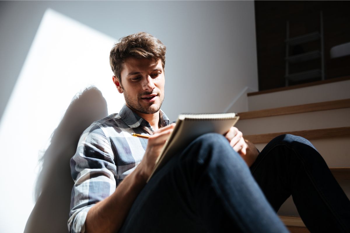 A man in monk mode, sitting on stairs, writing intently in a notebook, bathed in natural light.