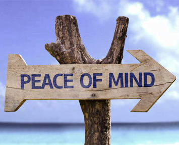 Wooden signpost with "peace of mind" written on it, pointing right, in front of a clear blue sky and ocean near a seaside store.