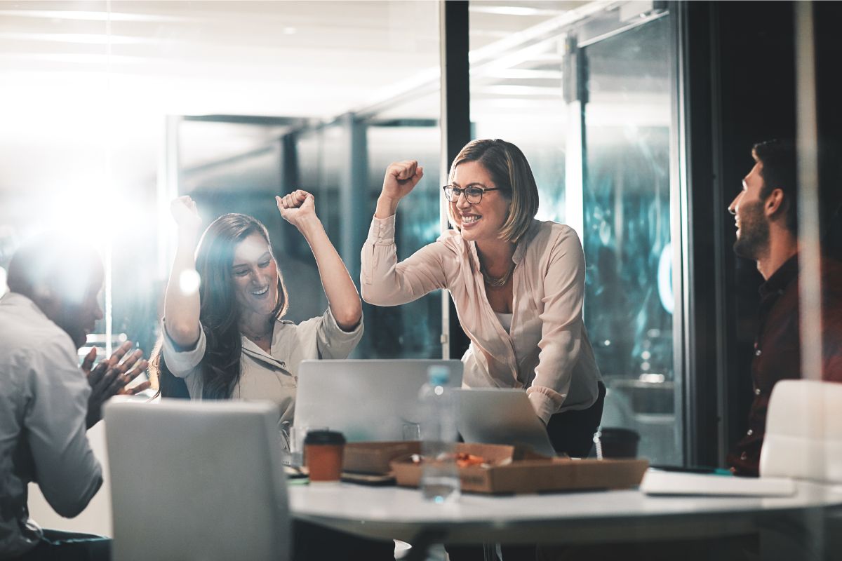 Two women celebrating with raised arms at a meeting with a male colleague clapping, in an Entrepreneurial Coaching session.