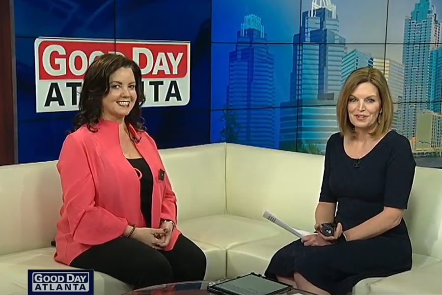 Two women seated on a white couch in a TV studio. A "Good Day Atlanta" sign is behind them. One is holding papers and a pen, the other is smiling, embodying the perfect media moment. Cityscape backdrop on right.