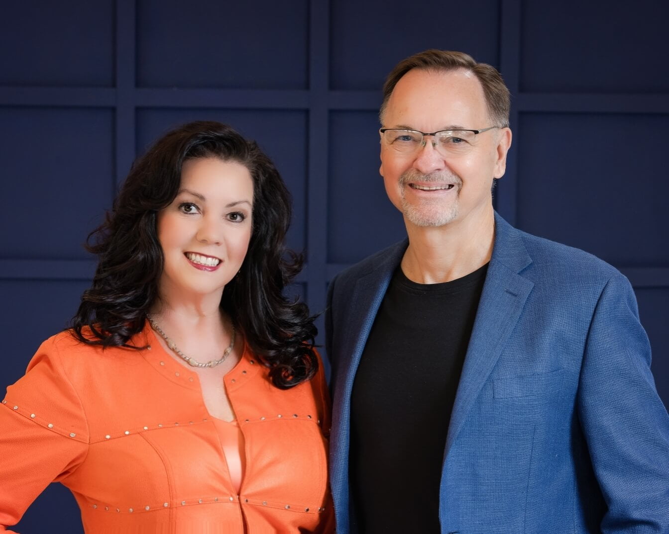 A woman in an orange top and a man in a blue blazer and black shirt smile while standing against a dark blue, square-patterned wall, bringing a touch of warmth to their stylish home.