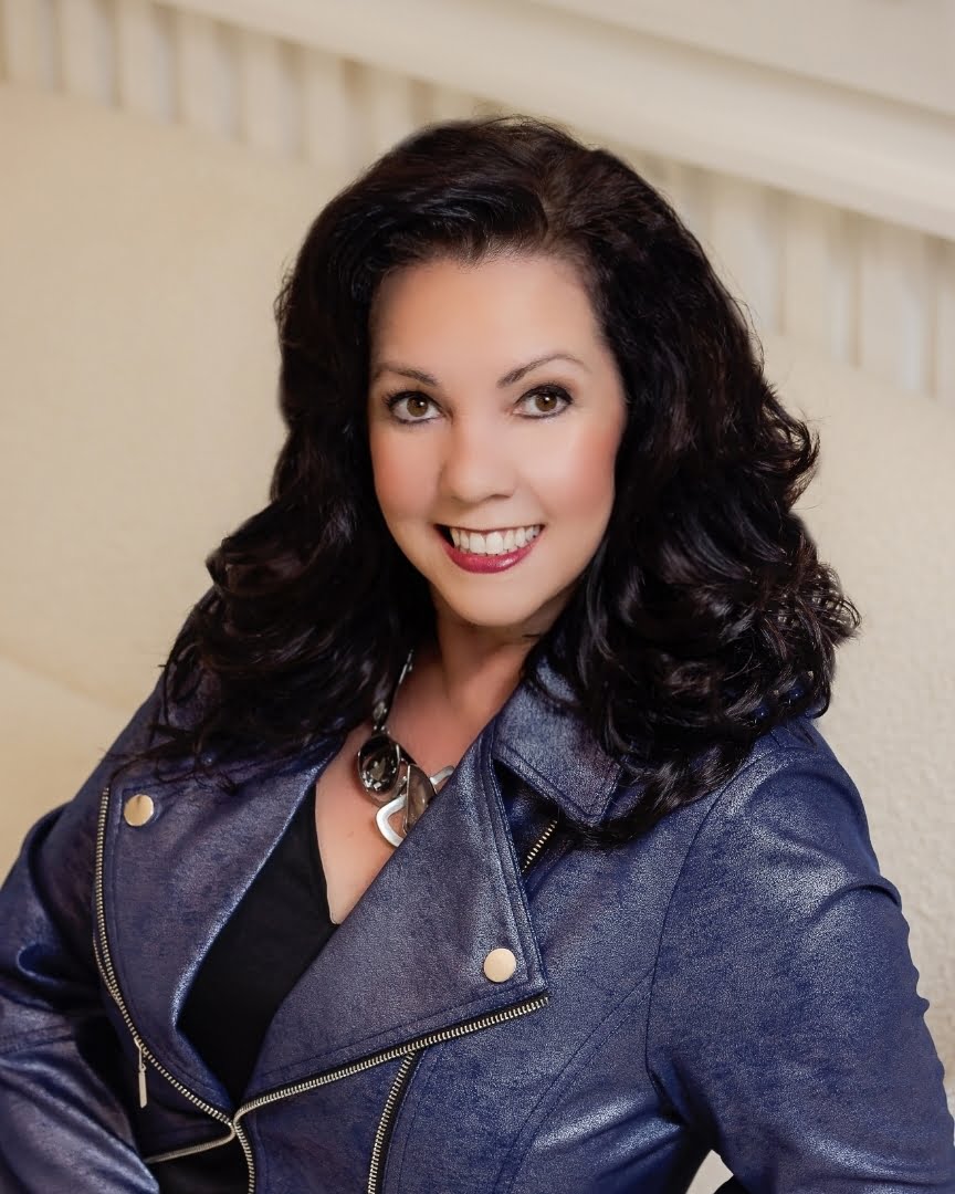 A woman with dark, curly hair smiles while seated on a light-colored couch. She is wearing a dark blue leather jacket and a statement necklace, embodying the style and warmth of our team featured in the About Us section.