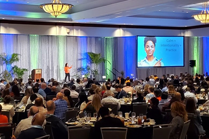 A person stands on stage addressing a seated audience in a conference room. A large screen displays a picture of another person and the text "Calm = Intentionality + Practice," fostering an atmosphere that feels almost as welcoming as home.