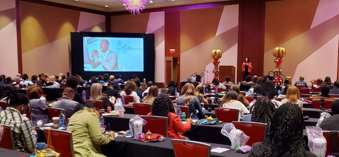 A conference room filled with seated attendees facing a presenter and a projected slide. The presenter, Angela Buttimer, stands near balloons and a screen displaying a person speaking.