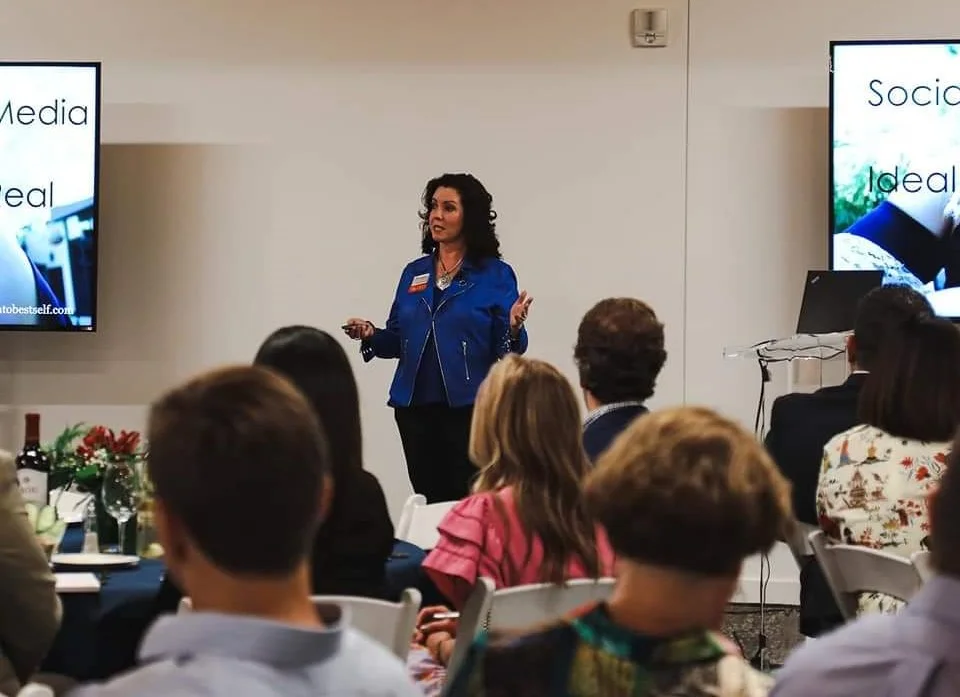 A woman delivers training on social media to an attentive audience seated in a room, with two screens displaying the keynotes in the background.