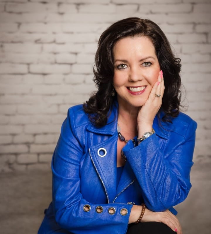 A woman with dark hair wearing a blue jacket poses contemplatively with her hand on her face in front of a white brick wall, evoking a sense of reflection often found in mental health therapy.