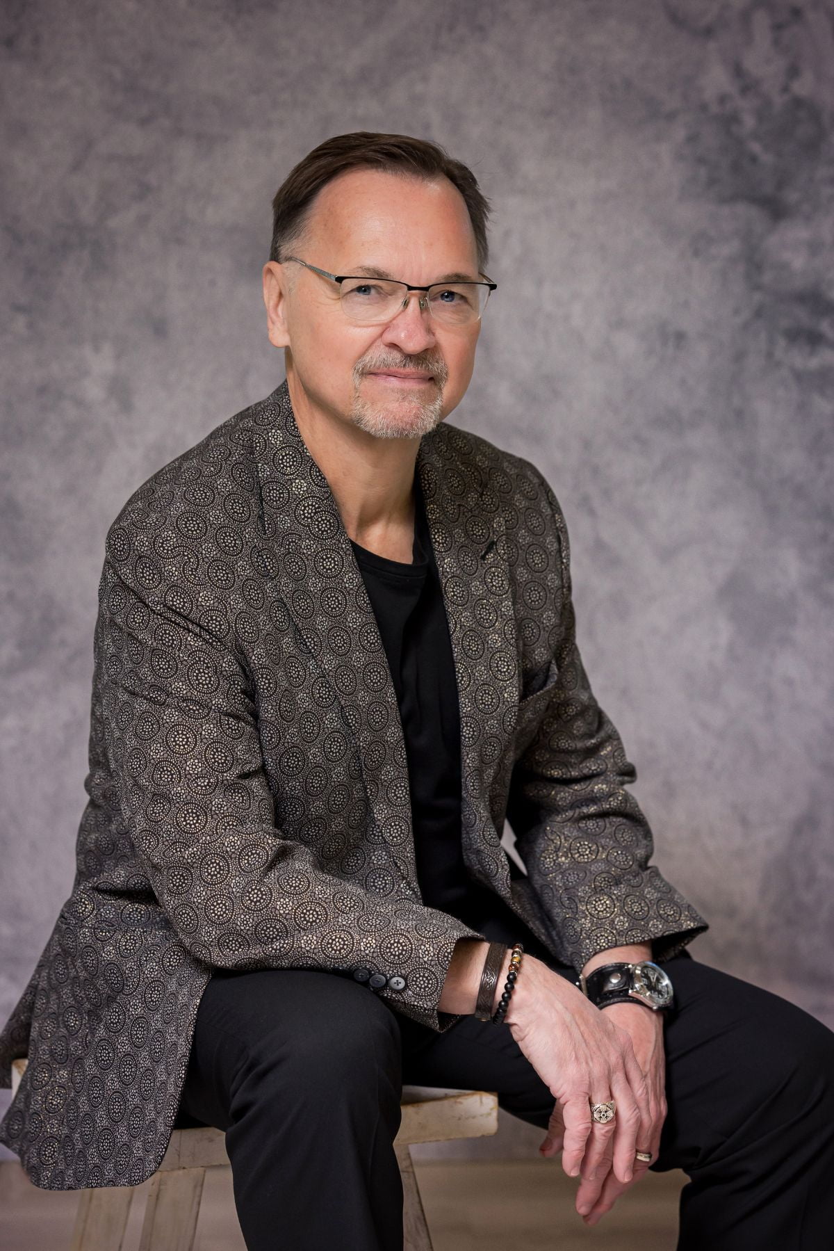 A man with glasses and a neat beard, wearing a patterned blazer and black shirt, sits on a stool against a gray studio backdrop, creating the perfect image for our About Us section.