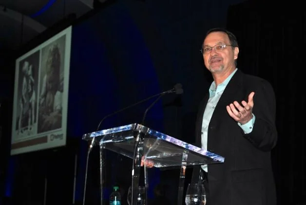 A man in a suit gestures while delivering keynotes at a podium on a stage with a projected image behind him.