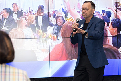 A man in a blue blazer is speaking into a microphone in front of a crowd at an event. The audience, feeling right at home, is seated at tables and appears engaged, with a large screen displaying the scene behind him.