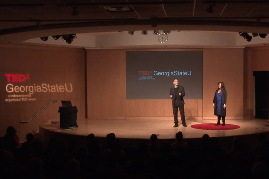 Two speakers stand on a stage under the title "TEDxGeorgiaStateU" projected behind them. The man on the left is speaking while the woman on the right listens intently. The audience, including several members of local media, is seated quietly.