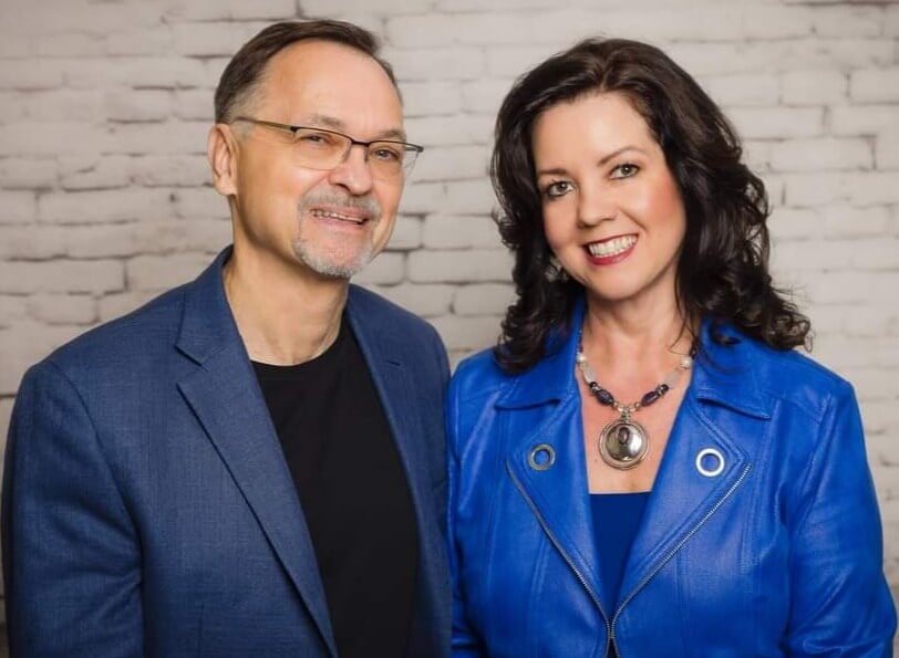 Angela And Dennis Buttimer, masterminds of Your Thought Leaders, pose smiling in front of a white brick background. Both are wearing blue jackets, and the woman has dark hair and a necklace.