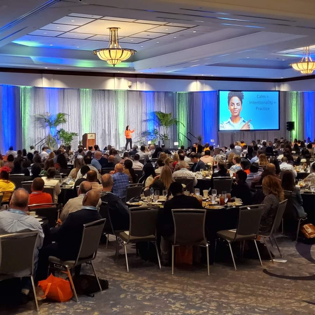 Angela Buttimer addresses a large seated audience in a conference room with a lit stage and projection screen displaying her presentation.