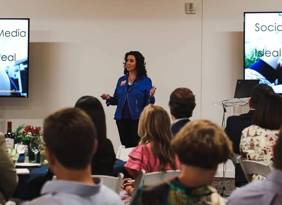 Angela Buttimer, dressed in a blue jacket, stands in front of a group of seated people, giving a presentation. Two screens display the text "Social Media Ideal.