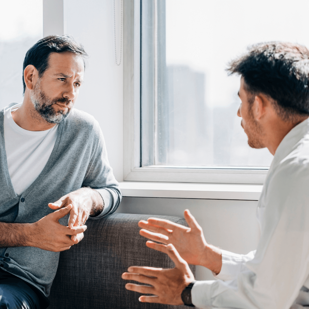 Two men are sitting and having a serious conversation about personal development in a brightly lit room near a window. One man gestures with his hands while the other listens intently, absorbing valuable life coaching insights.