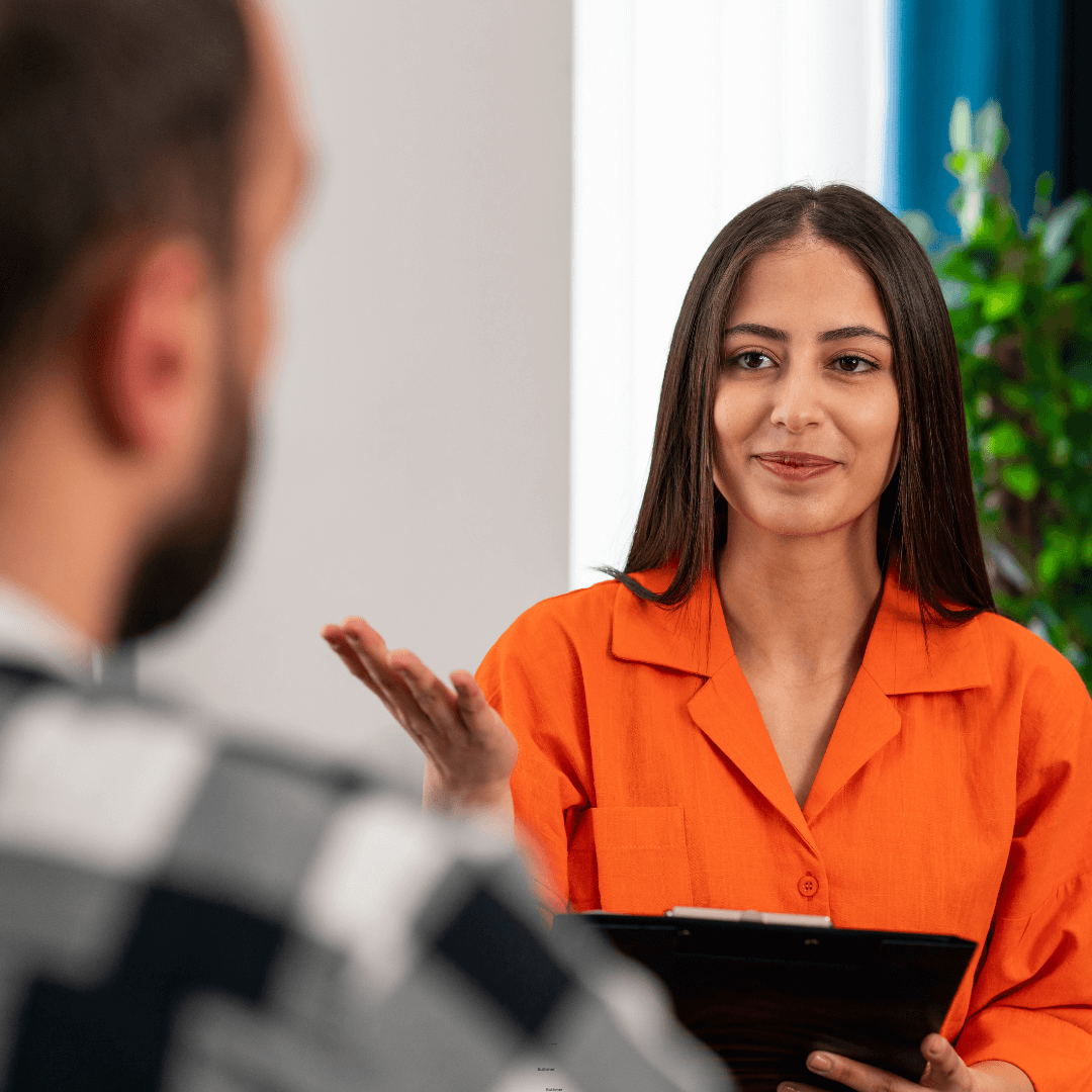 A woman wearing an orange shirt holds a tablet and gestures with her hand while discussing personal development with a man with short hair and a beard in focus in the foreground. A lush plant is visible in the background, adding to the atmosphere of self-improvement.