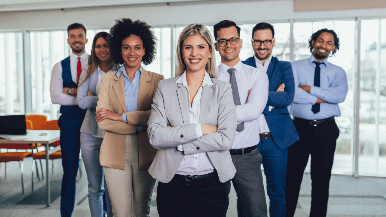 A group of seven people, dressed in professional attire, stands in an office with arms crossed, smiling at the camera, embodying strong leadership.