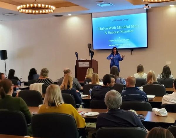 Angela Buttimer stands in front of an audience, presenting a slideshow titled "Thrive With Mindful Mojo: A Success Mindset" at a conference room. Attendees are seated at desks facing the presentation.