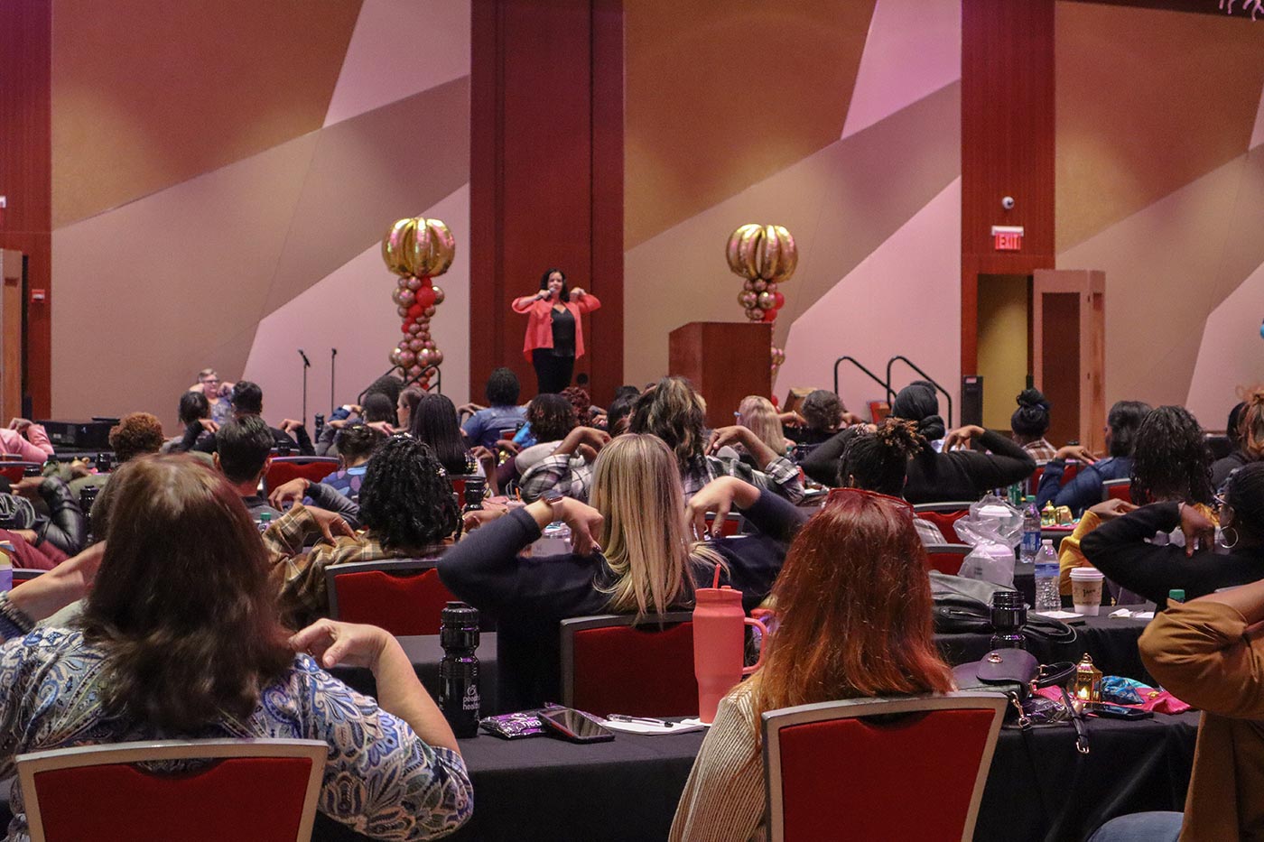 A speaker in a red blazer addresses a seated audience in a conference room. The attendees have their hands raised, participating in an interactive activity.