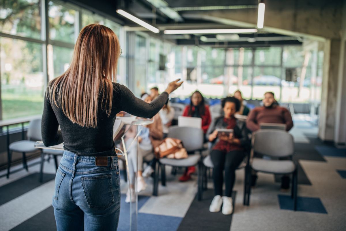 A woman stands at a podium addressing a small group of seated people in a modern, glass-walled room, discussing the benefits of coaching programs.