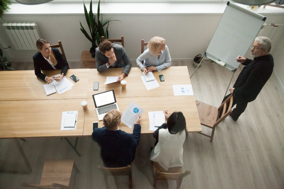 Six people in a meeting room, with five seated at a wooden table and one standing, writing on a whiteboard. Exhibiting strong teamwork, the group engages actively, with papers, pens, laptops, and coffee cups scattered on the table. The atmosphere highlights effective management practices.