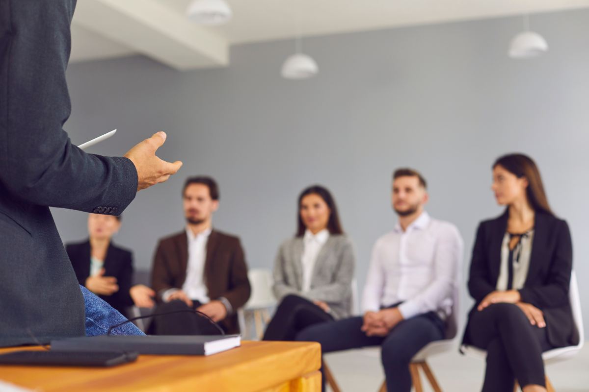 An expert in executive coaching is addressing a group of five seated individuals in a room with a grey wall.