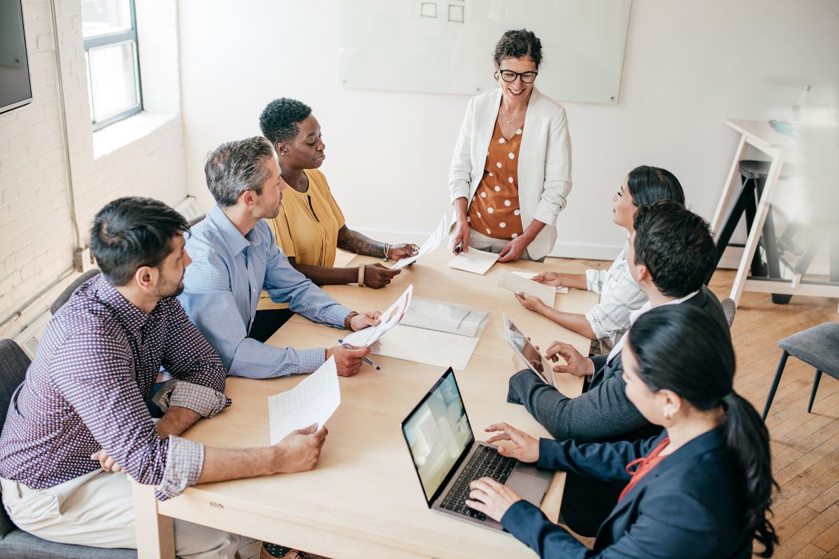 A group of six people is seated around a table in a meeting room, listening to an executive who is standing and speaking while holding papers. Some attendees are using laptops, while others have paper documents. This coaching session aims to enhance team performance and achieve organizational goals.