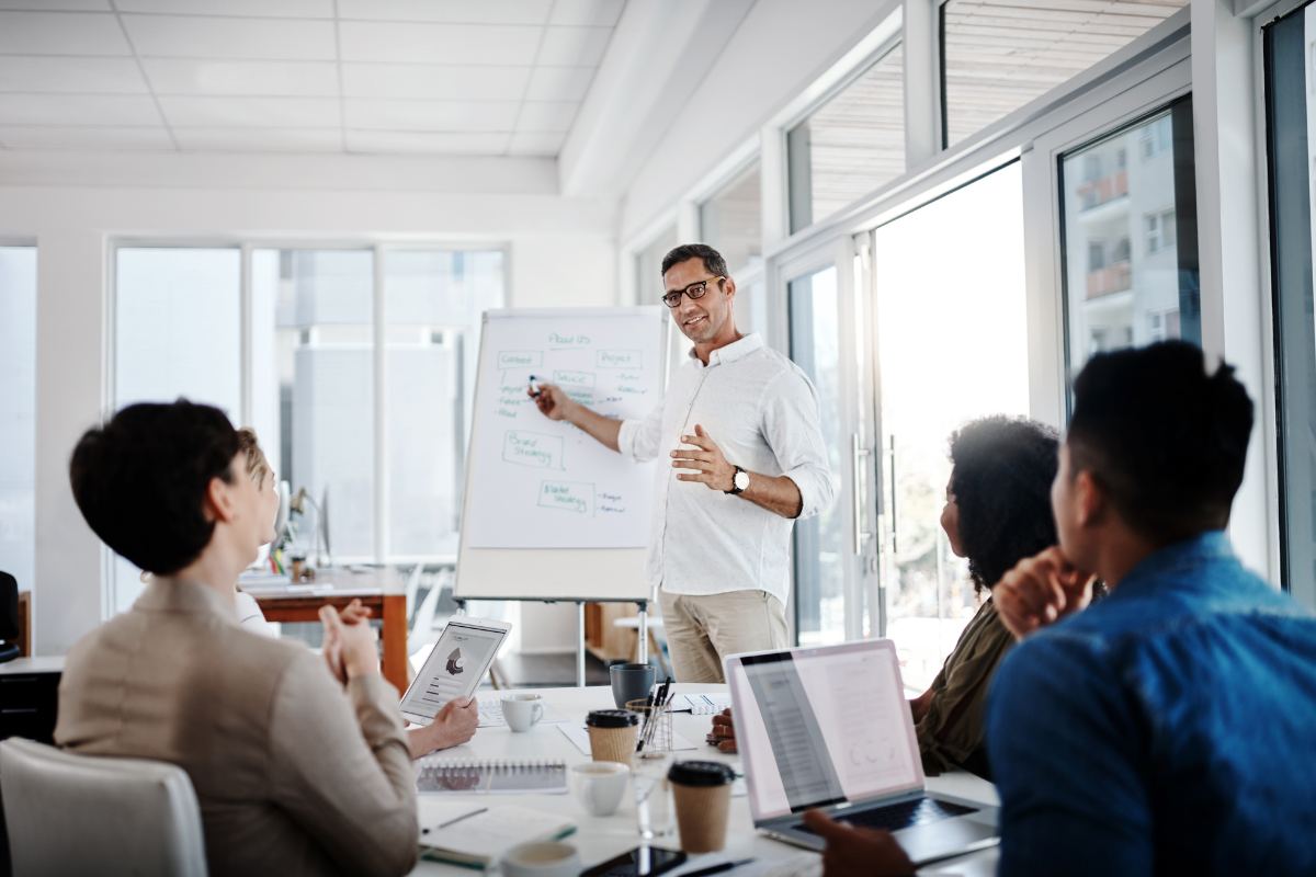 A man exudes leadership as he stands near a flip chart, presenting to a group of four seated individuals in a modern office setting. Some participants have laptops and notes in front of them.