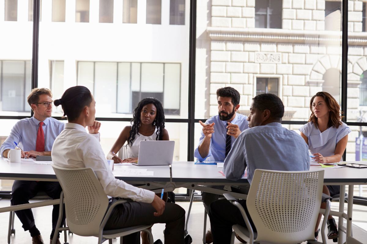 A diverse group of six people sit around a conference table, engaged in a leadership discussion. A laptop and papers are on the table. Large windows and an office building are visible in the background.