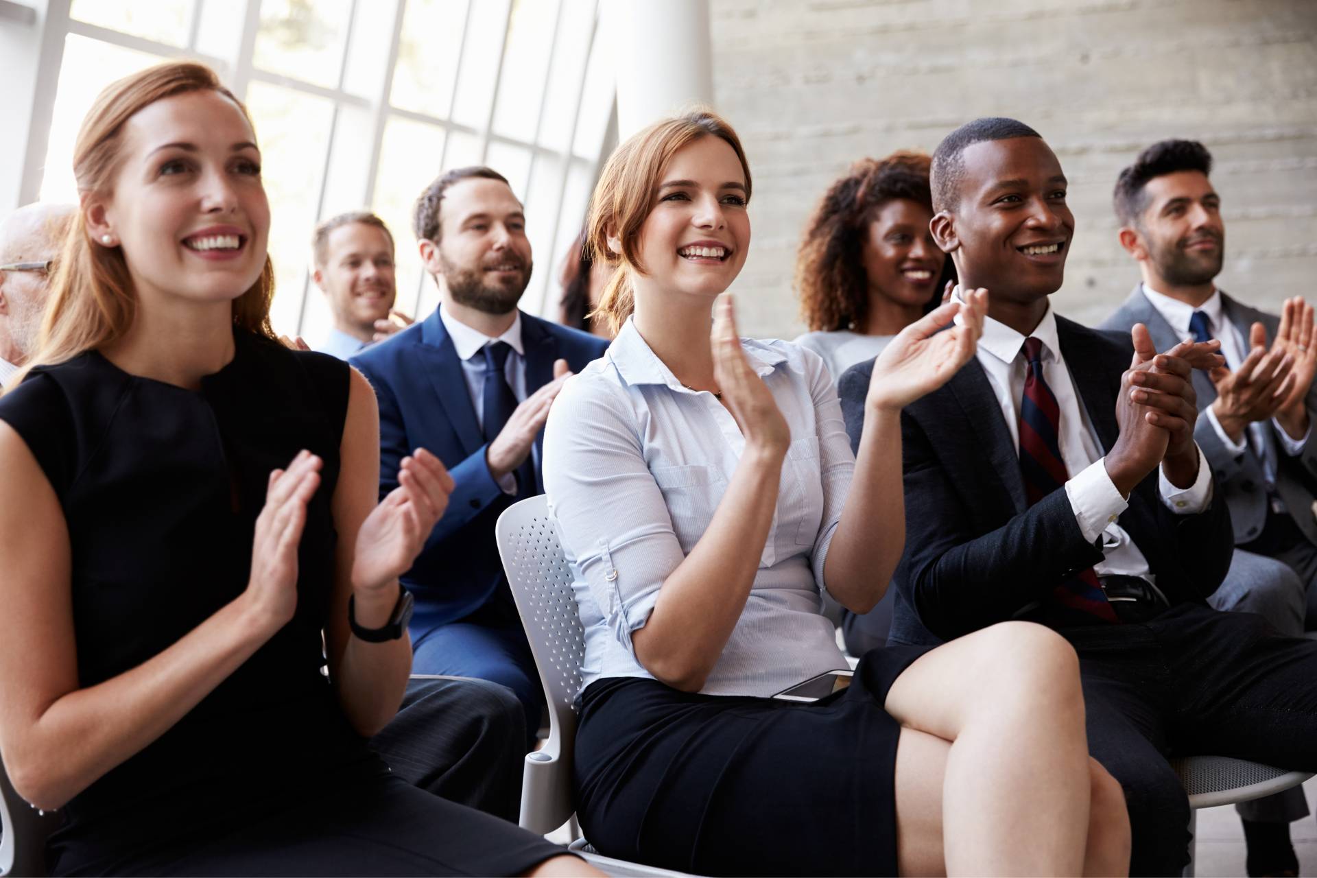 Audience members, dressed in business attire, sit in rows and applaud while smiling during a training session or keynote speech in a bright, modern room with large windows.