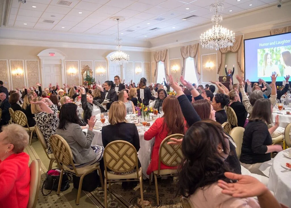 A large group of people seated at round tables in a banquet hall, many raising their hands. The room, set for a training session, has ornate decor with chandeliers and a presentation screen visible in the background.