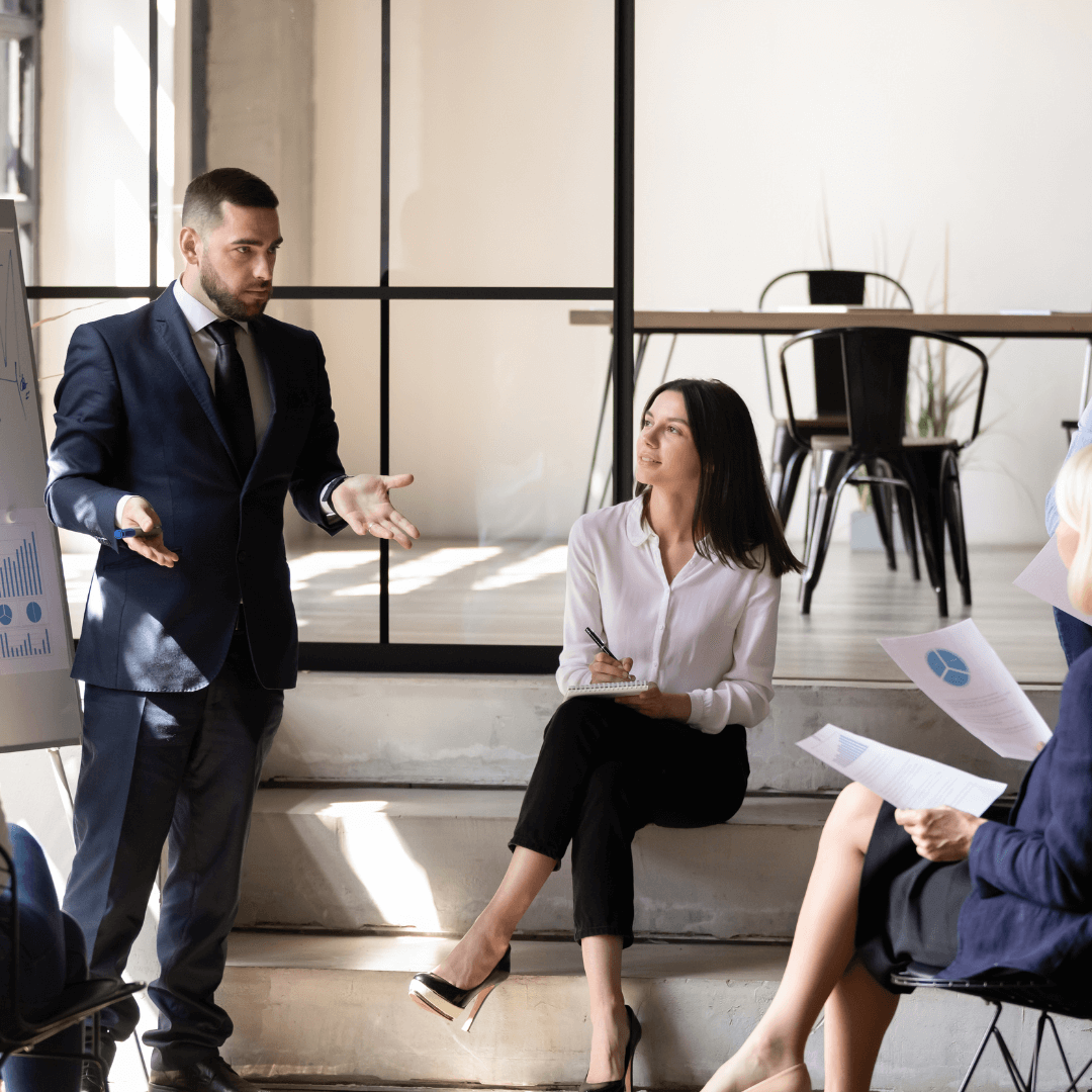 A man in a suit gives a presentation, gesturing with his hands, while three attentive masterminds listen and take notes. They are in a modern office space with large windows and minimal decor, creating the perfect environment for group coaching.