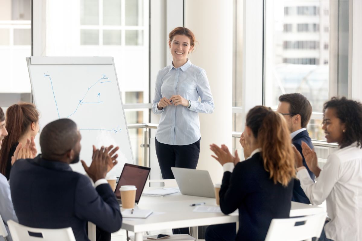 A woman stands in front of a whiteboard with a graph, smiling confidently, as a group of people, sitting around a table with laptops, applaud her leadership.