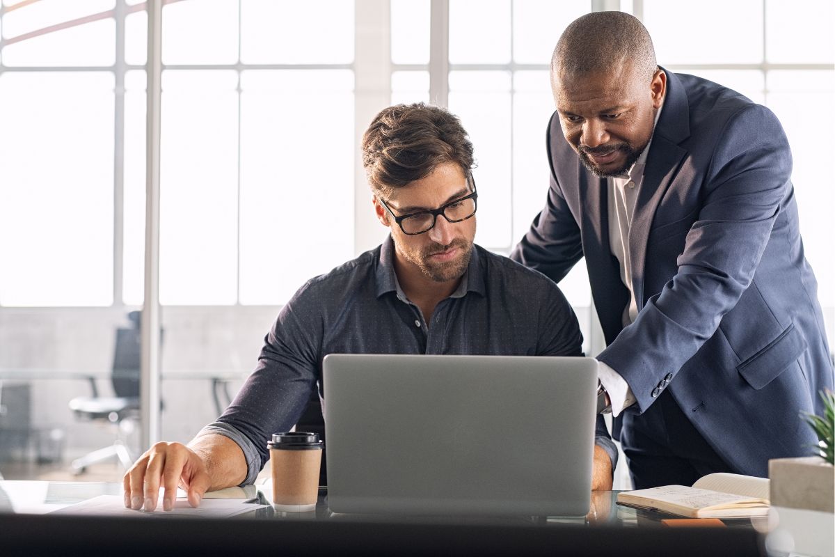 Two men in an office. One is seated, using a laptop, and the other is standing, pointing at the screen. There is a coffee cup and notebook on the desk. Both are focused and engaged in their work, demonstrating emotional intelligence in leadership through effective collaboration.