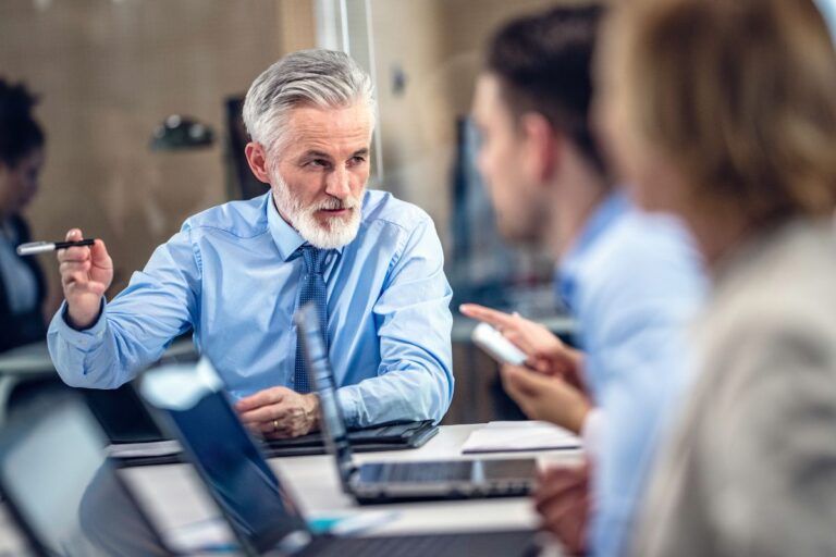 A man with gray hair and beard sits at a table, engaged in conversation with two other people. They appear to be in a business meeting, using laptops and documents, where the focus is on the importance of emotional intelligence in leadership.