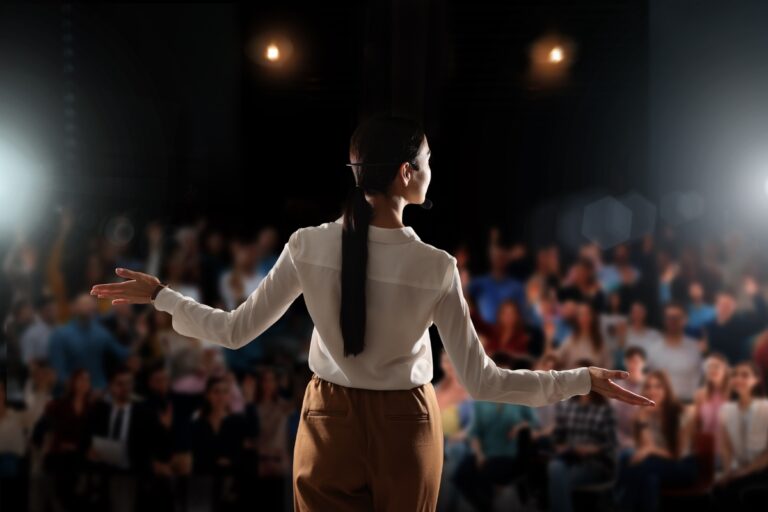 A person with long hair tied back, wearing a white shirt and brown pants, stands on stage as one of the inspirational keynote speakers, addressing a seated audience in a dimly lit room with bright lights shining from behind.