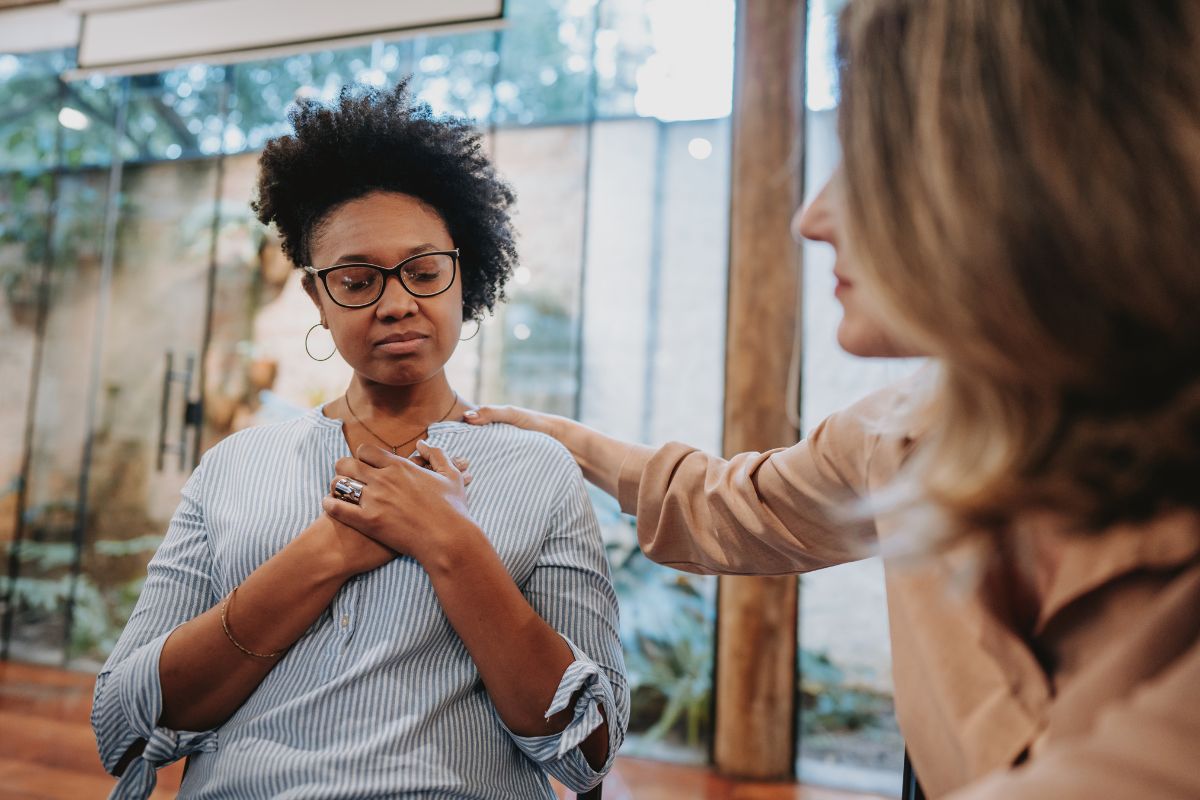 A woman with glasses holds her hands over her chest while another woman places a comforting hand on her shoulder. They are in a room with large windows, demonstrating emotional intelligence in leadership.