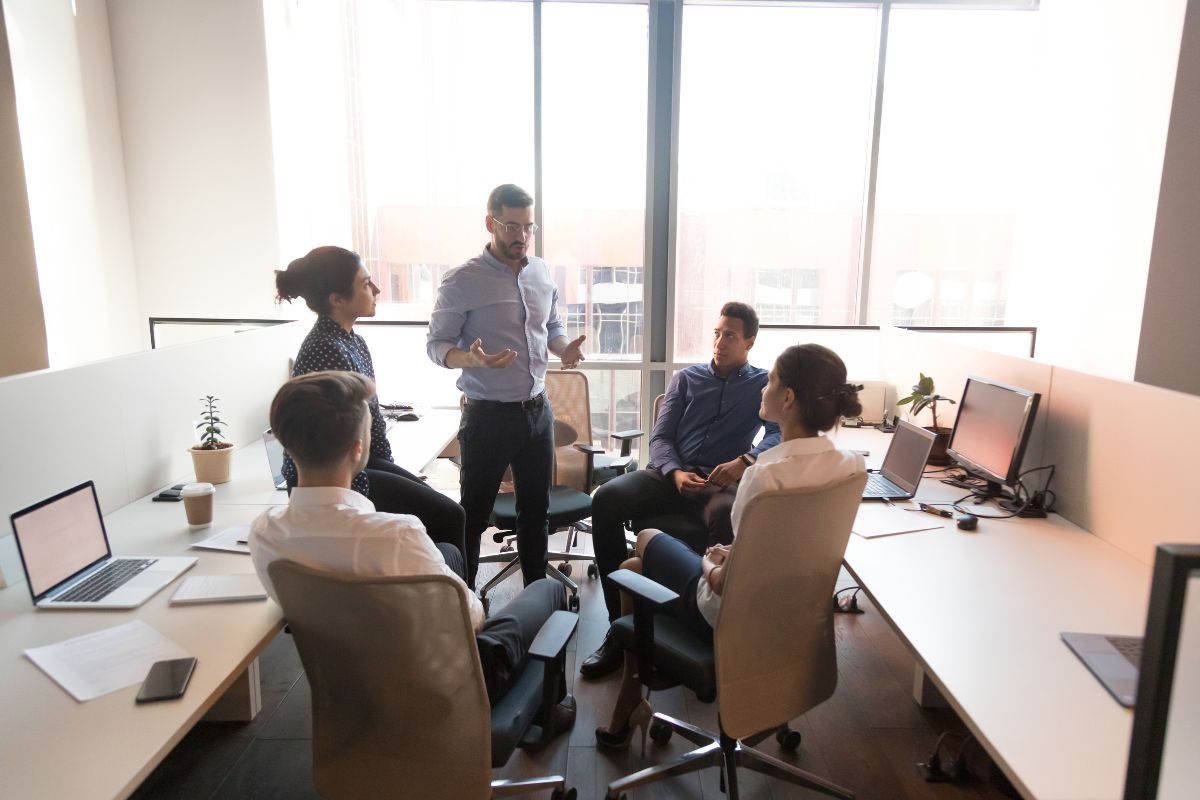 Several people are gathered in a modern office space, engaging in a discussion that highlights the role of emotional intelligence in leadership. They are seated around workstations with laptops, papers, and office supplies visible. Large windows provide a bright backdrop to their collaborative efforts.