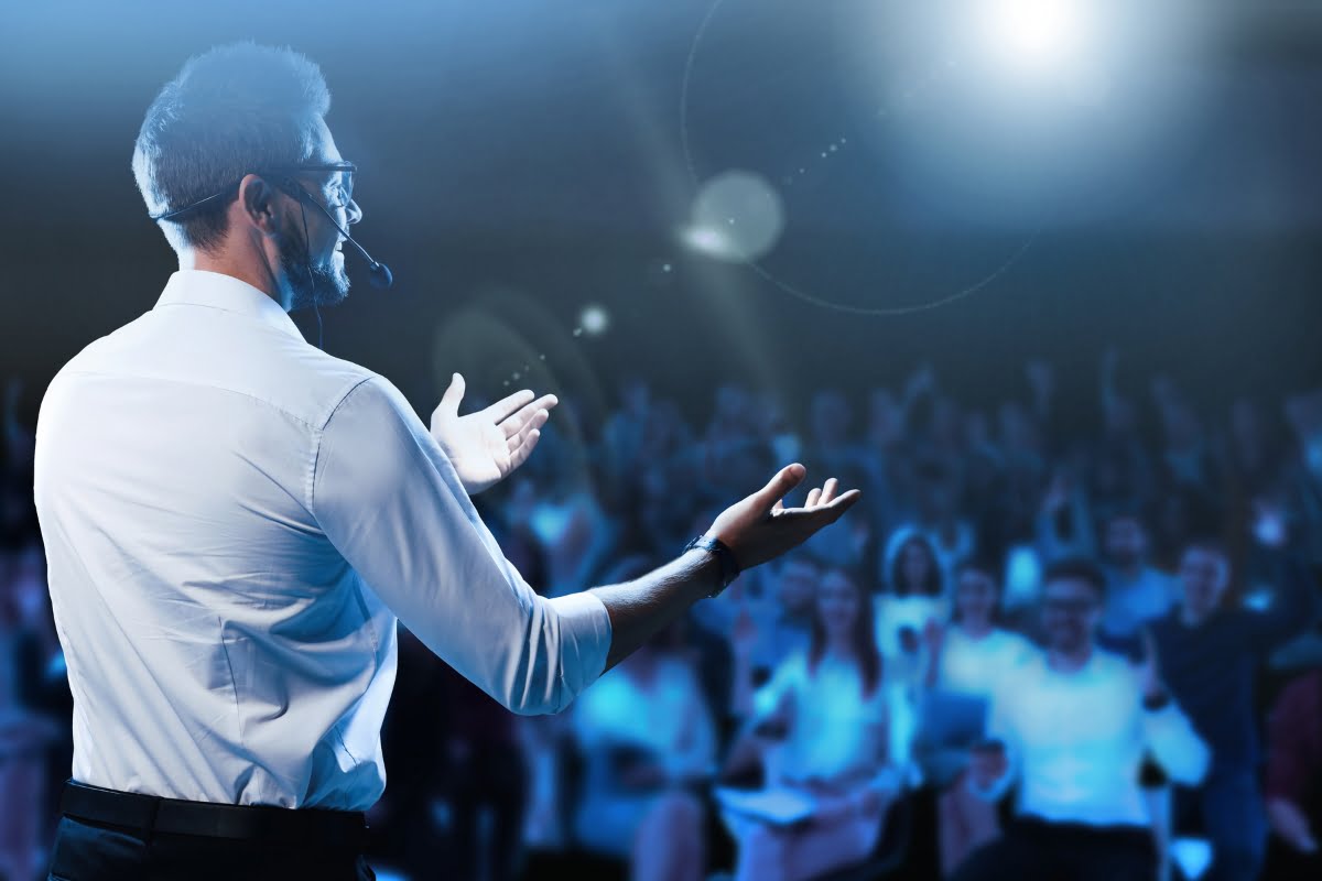 A man in a white shirt and headset, potentially ideal when choosing a keynote speaker, speaks energetically to a large, blurred audience under bright stage lighting.